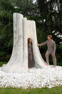 A boy and a girl are playing hide and seek in a white carrara marble sculpture resembling a tree. They are both wearing a double-twisted cotton pajama with nog-yellow and dark blue stripes.