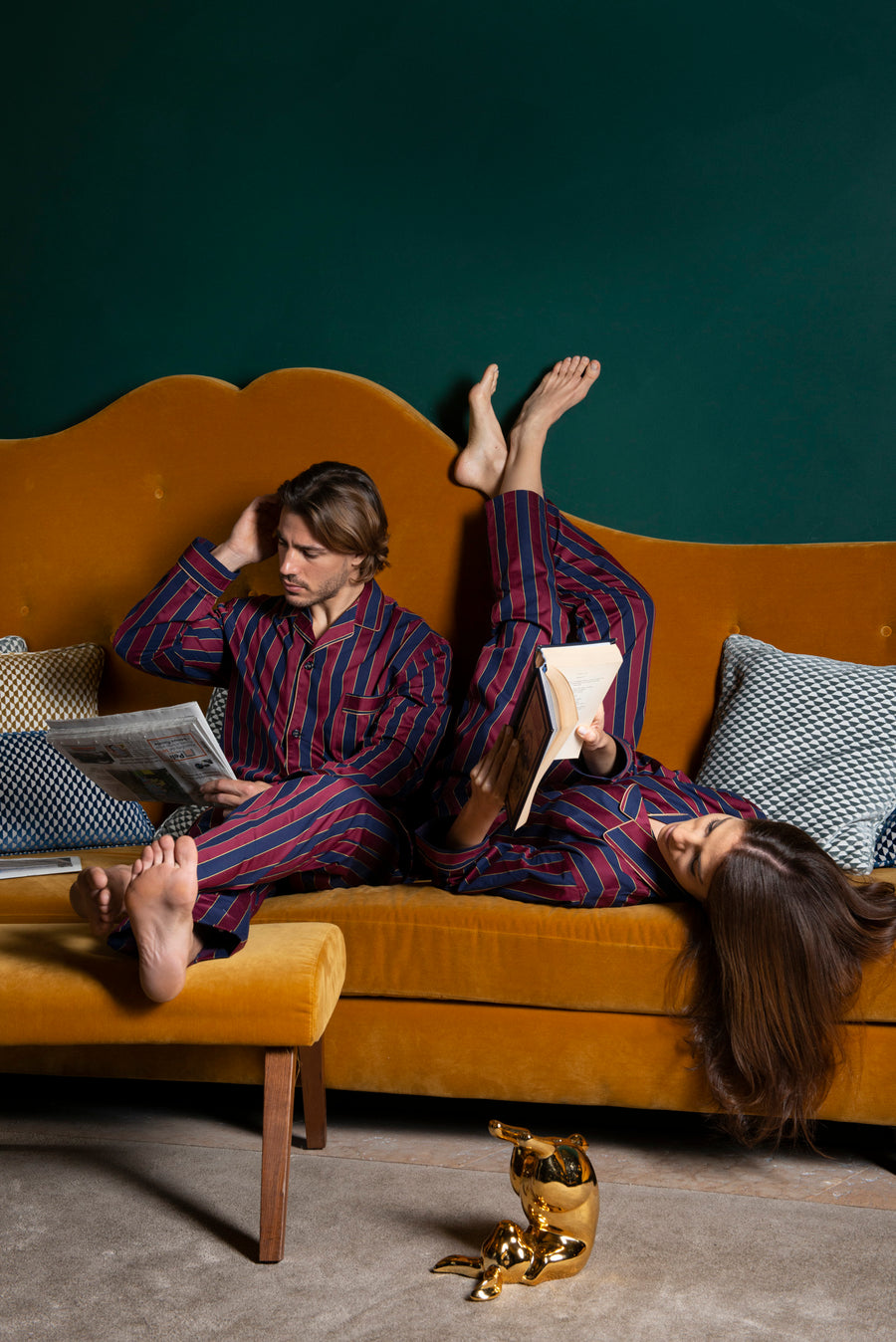 A boy and a girl are sitting on a yellow couch. He is reading a newspaper, she is reading Shakespeare while sitting in reverse. A golden rabbit is rolling on his back on the floor in front of them. They are both wearing a cotton satin macro striped pajama in burgundy and dark blue.