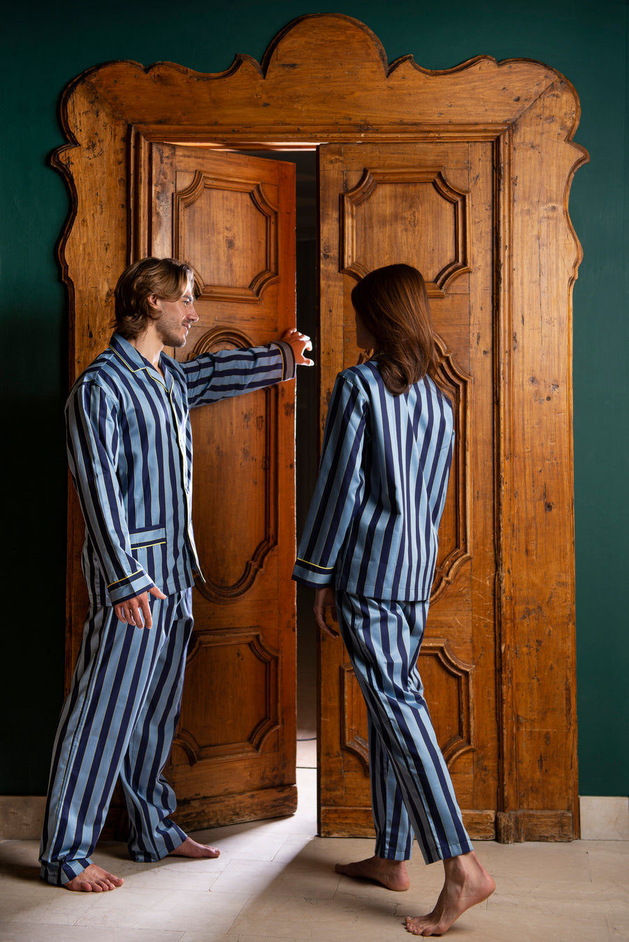 A boy and a girl are in front of a wooden antique gate. He is opening the gate and inviting the girl to enter. They are both wearing a cotton satin macro striped pajama in avio and dark blue.